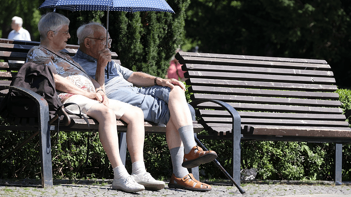 Älteres Paar sitzt an einem sonnigen Tag im Park auf einer Bank unter einem Schirm – Symbolbild für Hitzeschutz.